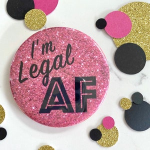 May include: Pink glitter button with black text that reads "I'm Legal AF". The button is surrounded by pink, black, and gold confetti.