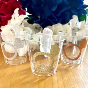 May include: Three clear plastic shot glasses with gold, silver, and copper ring designs. The rings have glitter accents.
