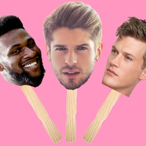 May include: Three cardboard cutouts of men's faces on wooden sticks. The men have different hairstyles and skin tones. The background is pink.