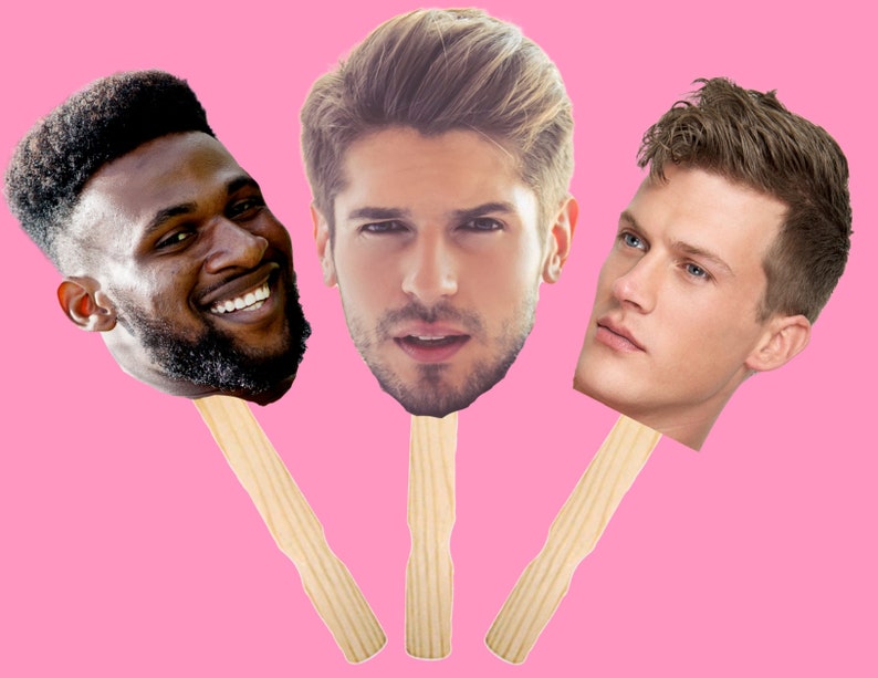 May include: Three cardboard cutouts of men's faces on wooden sticks. The men have different hairstyles and skin tones. The background is pink.