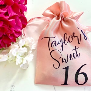 May include: A pink satin drawstring bag with black text that reads "Taylor's Sweet 16".