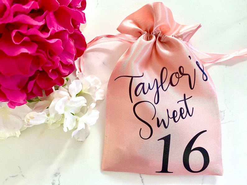 May include: A pink satin drawstring bag with black text that reads "Taylor's Sweet 16".