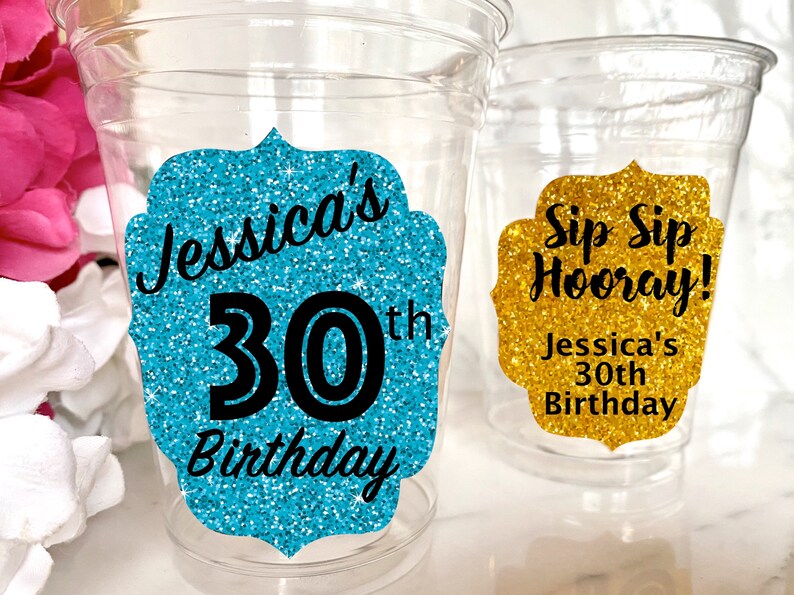 May include: Two clear plastic cups with glitter birthday party decorations. One cup has a turquoise glitter sticker with the text "Jessica's 30th Birthday". The other cup has a gold glitter sticker with the text "Sip Sip Hooray! Jessica's 30th Birthday".
