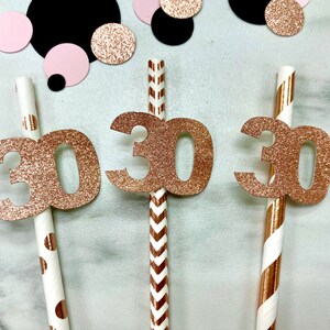 May include: Three paper straws with rose gold glitter "30" toppers, surrounded by pink, black, and rose gold confetti. The straws have different patterns: polka dots, chevrons, and stripes. A festive decoration for a 30th birthday celebration.
