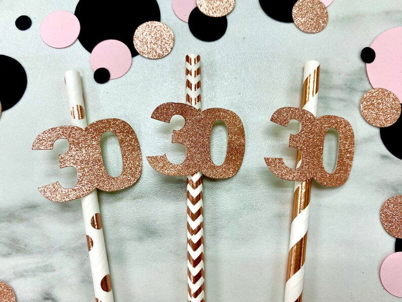May include: Three paper straws with rose gold glitter "30" toppers, surrounded by pink, black, and rose gold confetti. The straws have different patterns: polka dots, chevrons, and stripes. A festive decoration for a 30th birthday celebration.