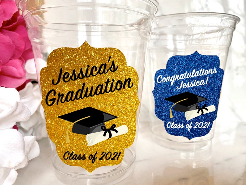 Puede incluir: Dos vasos de pl&aacute;stico transparentes con decoraciones para una fiesta de graduaci&oacute;n. Un vaso tiene una pegatina de purpurina dorada con el texto "Jessica's Graduation Class of 2021" y un gorro de graduaci&oacute;n negro y un diploma. El otro vaso tiene una pegatina de purpurina azul con el texto "Congratulations Jessica! Class of 2021" y un gorro de graduaci&oacute;n negro y un diploma.