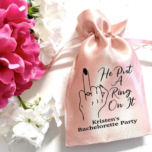 May include: Pink satin bag with the text "He Put A Ring On It" and "Kristen's Bachelorette Party" printed on it. The bag features a black and white illustration of a hand with a ring on the finger.