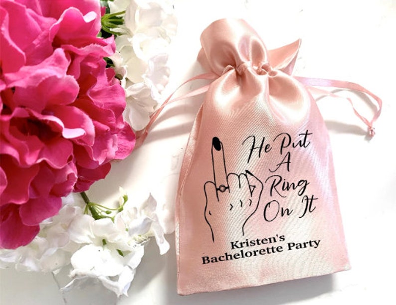 May include: Pink satin bag with the text "He Put A Ring On It" and "Kristen's Bachelorette Party" printed on it. The bag features a black and white illustration of a hand with a ring on the finger.