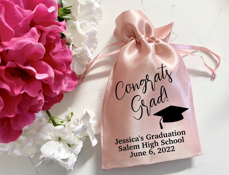 Puede incluir: Bolsa de cord&oacute;n de sat&eacute;n rosa con texto negro que dice "Congrats Grad!" y un gr&aacute;fico de gorro de graduaci&oacute;n negro. La bolsa tambi&eacute;n tiene el texto "Jessica's Graduation Salem High School June 6, 2022".