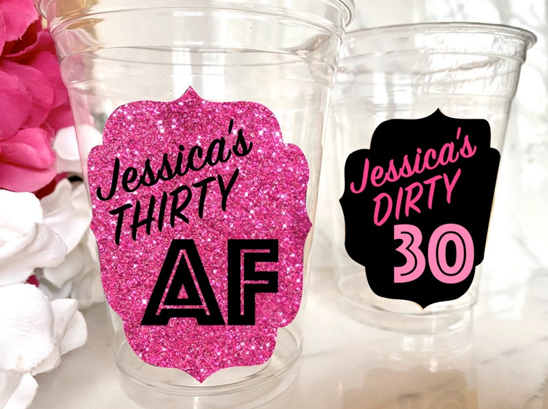 May include: Two clear plastic cups with pink and black glitter stickers. The sticker on the left cup says "Jessica's Thirty AF" in black text on a pink glitter background. The sticker on the right cup says "Jessica's Dirty 30" in pink text on a black background.