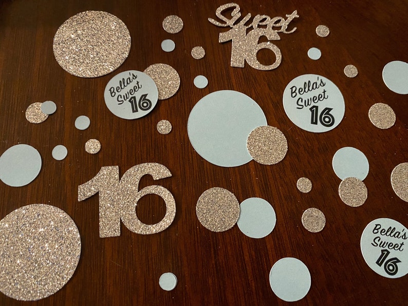 May include: Gold glitter confetti with white and gold circles, and two large gold glitter numbers 16. There are also three circles with the text "Bella's Sweet 16" in black.