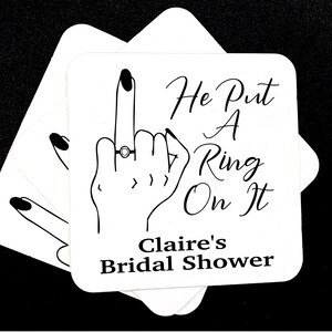 May include: White cardstock coaster with a black line drawing of a hand with a ring on the middle finger. The text reads "He Put A Ring On It" and "Claire's Bridal Shower".