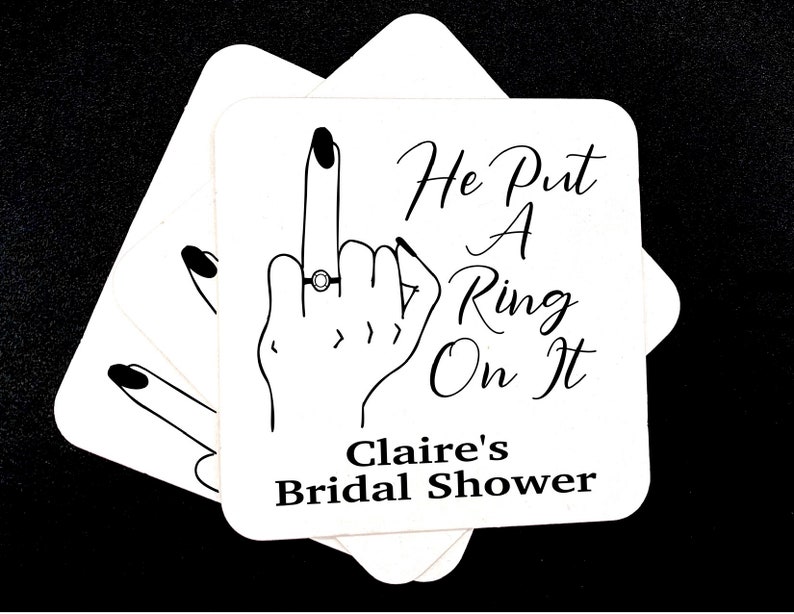 May include: White cardstock coaster with a black line drawing of a hand with a ring on the middle finger. The text reads "He Put A Ring On It" and "Claire's Bridal Shower".
