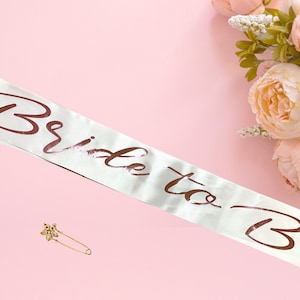 May include: White satin sash with the text "Bride to Be" in rose gold lettering. A gold safety pin is included.