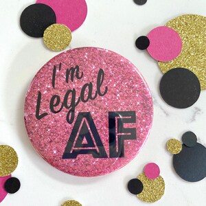 May include: Pink glitter button with the text "I'm Legal AF" in black lettering.