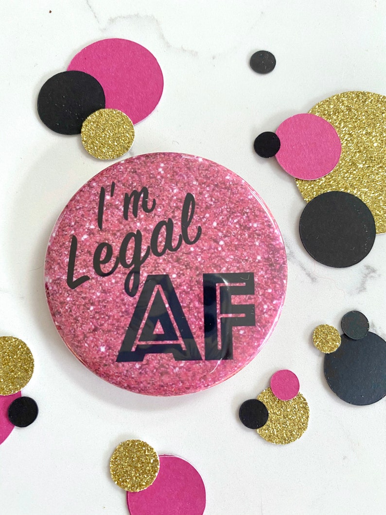 May include: Pink glitter button with the text "I'm Legal AF" in black lettering.