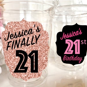 May include: Two clear plastic cups with personalized 21st birthday party decorations. One cup has a rose gold glitter background with black text that says "Jessica's FINALLY 21". The other cup has a black background with pink text that says "Jessica's 21st Birthday".
