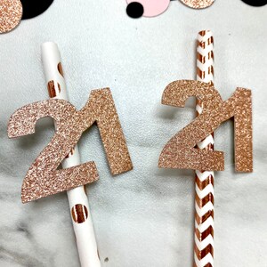 May include: Two gold glitter straws with the number 21 attached to them. One straw has white polka dots and the other has a chevron pattern.