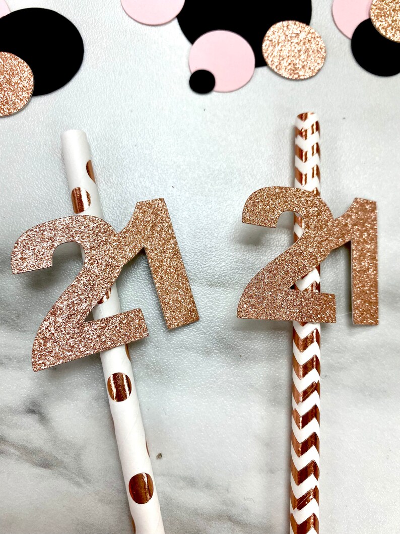 May include: Two gold glitter straws with the number 21 attached to them. One straw has white polka dots and the other has a chevron pattern.