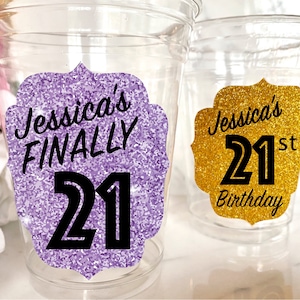 May include: Two clear plastic cups with glitter-covered labels. The purple label says "Jessica's FINALLY 21" and the gold label says "Jessica's 21st Birthday".