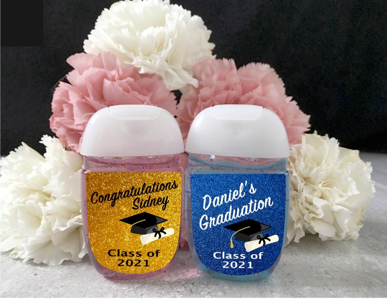 May include: Two small bottles of hand sanitizer with graduation themed labels. The bottle on the left has a gold glitter background with the text 'Congratulations Sidney Class of 2021'. The bottle on the right has a blue glitter background with the text 'Daniel's Graduation Class of 2021'.