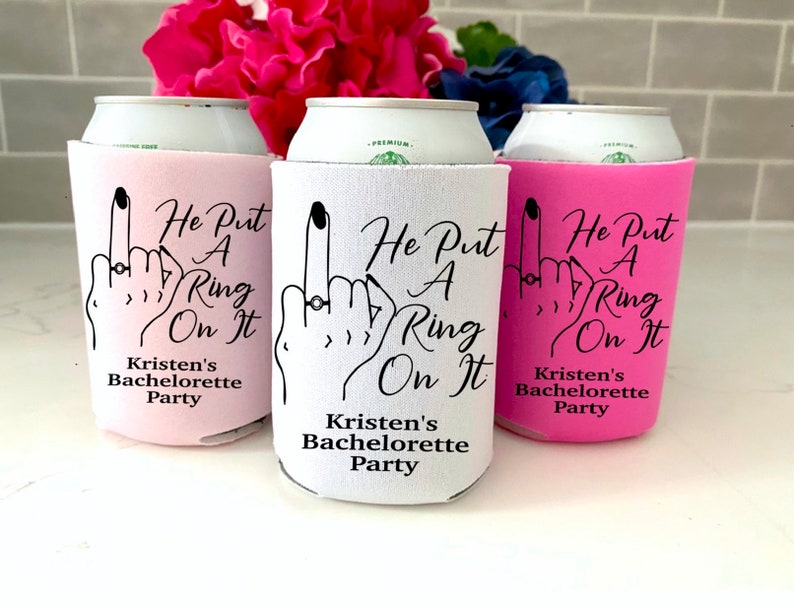 May include: Three pink, white, and black can coolers with the text "He Put A Ring On It" and "Kristen's Bachelorette Party".