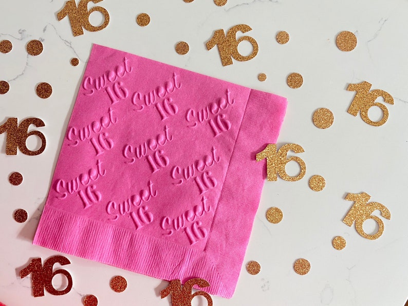 May include: Pink napkins with the words "Sweet 16" embossed in white. The napkins are surrounded by gold glitter confetti with the number 16.
