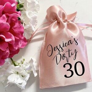 May include: A pink satin drawstring bag with black text that reads "Jessica's Dirty 30".