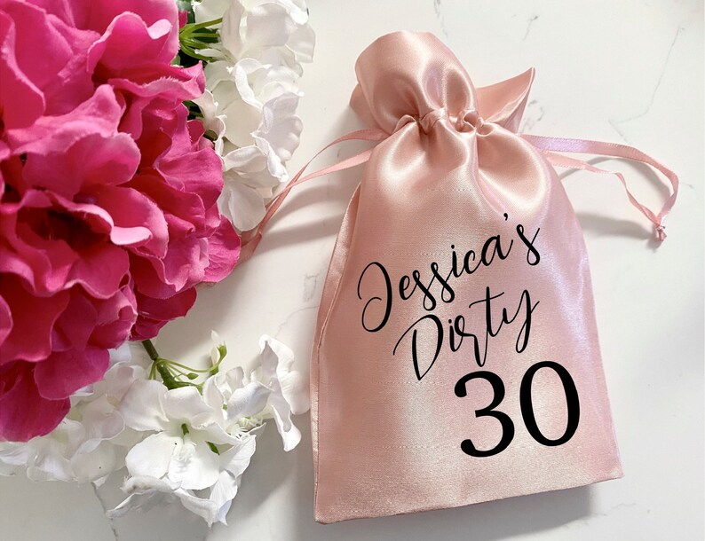 May include: A pink satin drawstring bag with black text that reads "Jessica's Dirty 30".