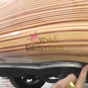 May include: A close-up of a car's rear bumper covered in a wood-grain vinyl wrap. The wrap has a light brown and reddish-brown color scheme. The text "JDM BESTBOY" is visible on the wrap.