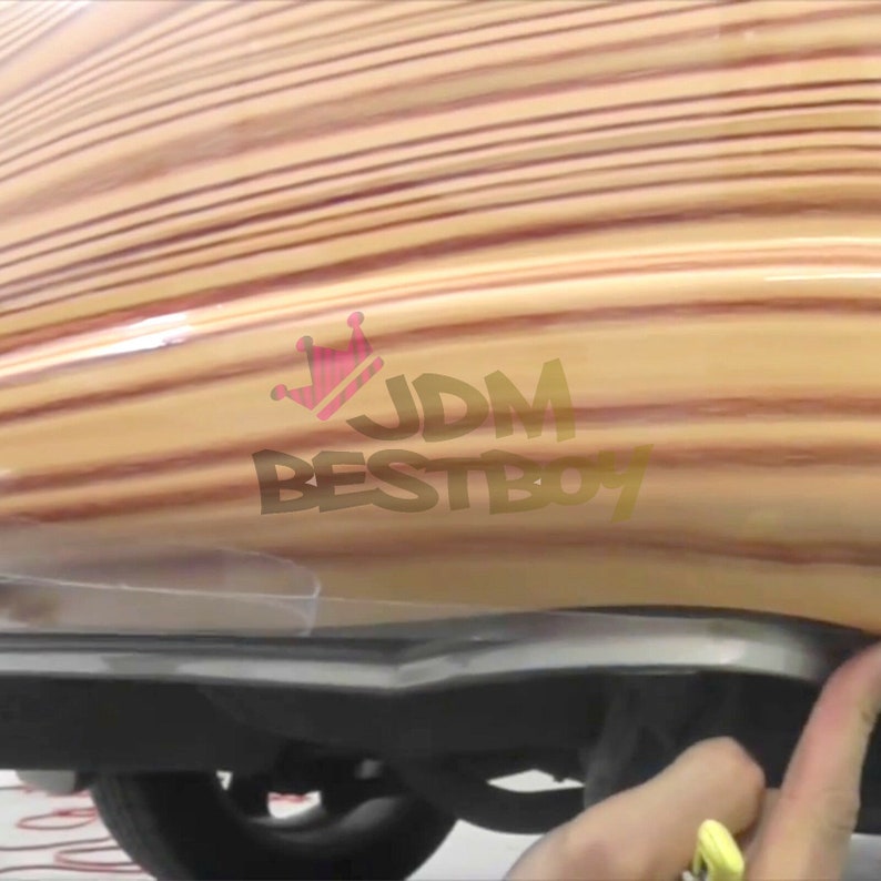 May include: A close-up of a car's rear bumper covered in a wood-grain vinyl wrap. The wrap has a light brown and reddish-brown color scheme. The text "JDM BESTBOY" is visible on the wrap.