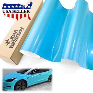 May include: A roll of glossy light blue vinyl wrap film next to a cardboard box labeled JDM BESTBOY with a USA map graphic. Below, a light blue sedan is shown with the same glossy finish, featuring black wheels and black window trim details.