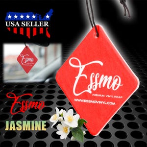 May include: Red diamond-shaped car air freshener with the text "Esso Premium Vinyl Wrap www.essmovinyl.com" in white. The air freshener is hanging from a black string. A white jasmine flower is in the foreground.
