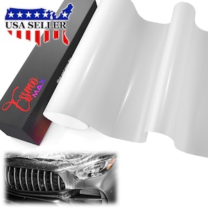 May include: A roll of glossy white vinyl wrap film positioned next to a black rectangular box labeled Essmo Max. The box features a USA Seller graphic. Below, a partial view of a silver car front bumper and headlight shows the film applied.