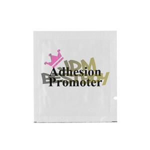 May include: A small, white packet with the words "Adhesion Promoter" in black and gold lettering. A pink crown graphic is above the text. The packet is sealed on all sides.