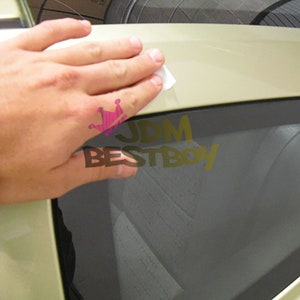 May include: A hand applying a white cleaning wipe to a light-colored car surface. The image includes the text "JDM BESTBOY" in gold lettering with a pink crown graphic.