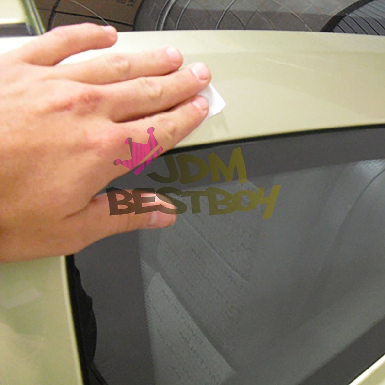 May include: A hand applying a white cleaning wipe to a light-colored car surface. The image includes the text "JDM BESTBOY" in gold lettering with a pink crown graphic.