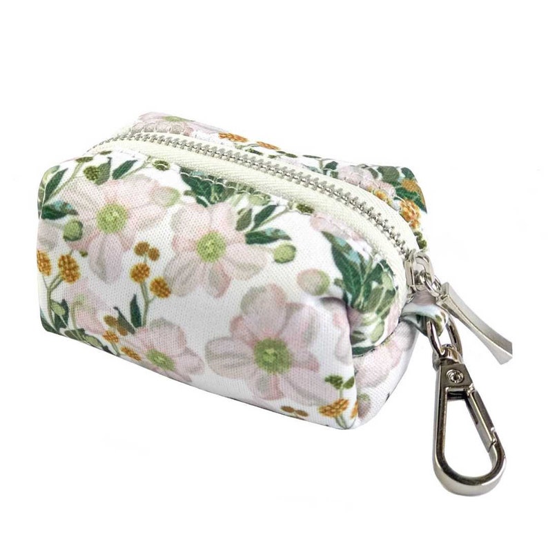 May include: A white zippered pouch with a floral print featuring pink flowers, green leaves, and yellow accents. The pouch has a silver metal clip.