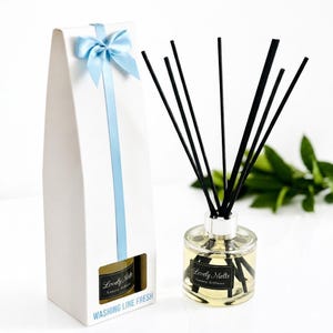 May include: A reed diffuser with a clear glass bottle, black reeds, and a white gift box. The bottle and box feature a black label with the text "Lovely Melts Luxury Diffuser". The box has a light blue ribbon and bow, with "WASHING LINE FRESH" printed in blue.