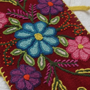 Puede incluir: Textil rectangular bordado con fondo rojo oscuro. El diseño presenta flores coloridas en tonos rosa, azul, morado y amarillo, con hojas y tallos verdes. Se utiliza hilo amarillo para el borde y las presillas para colgar.