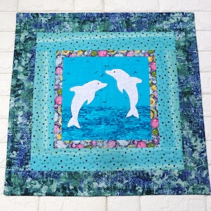 May include: A blue and green patchwork quilt with a square center featuring two white dolphin silhouettes swimming in a blue ocean. The quilt is framed with multiple layers of fabric with polka dots and a blue and green swirl pattern.