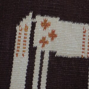 May include: Woven textile rug with a geometric design featuring a stylized llama in cream and brown. The llama has orange cross-shaped accents and is set against a striped background. The rug is made of a textured material.