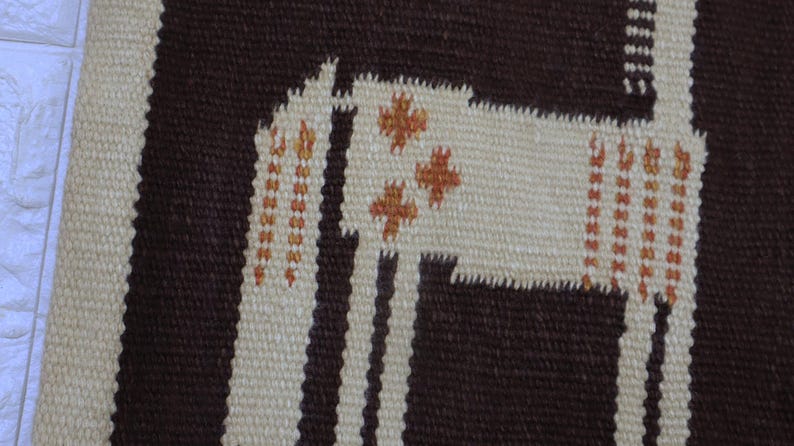 May include: Woven textile rug with a geometric design featuring a stylized llama in cream and brown. The llama has orange cross-shaped accents and is set against a striped background. The rug is made of a textured material.
