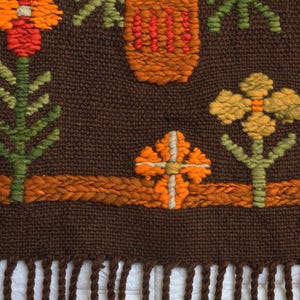 Op de afbeelding: Een bruine geweven wandhanger met een franje rand. De hanger heeft een geometrisch ontwerp met oranje, gele en groene bloemen.