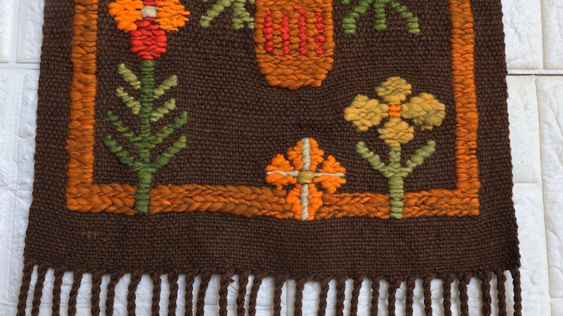 Op de afbeelding: Een bruine geweven wandhanger met een franje rand. De hanger heeft een geometrisch ontwerp met oranje, gele en groene bloemen.