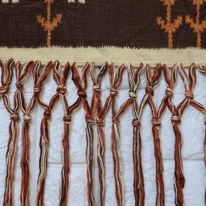 May include: A woven wall hanging with a brown and beige geometric pattern at the top. The bottom features knotted fringe in shades of brown and beige. The textile is likely handmade.