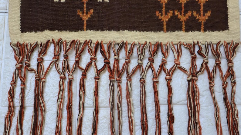 May include: A woven wall hanging with a brown and beige geometric pattern at the top. The bottom features knotted fringe in shades of brown and beige. The textile is likely handmade.