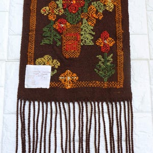 Op de afbeelding: Een bruine geweven wandhanger met een bloemenontwerp met een vaas met oranje, rode en gele bloemen. De hanger heeft een franje rand met bruine, gele en rode accenten.