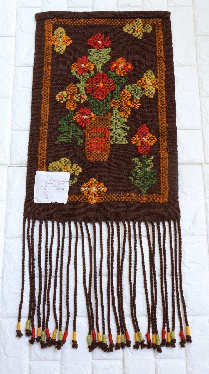 Op de afbeelding: Een bruine geweven wandhanger met een bloemenontwerp met een vaas met oranje, rode en gele bloemen. De hanger heeft een franje rand met bruine, gele en rode accenten.