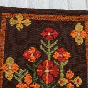 Op de afbeelding: Een bruin vloerkleed met een bloemenpatroon. Het vloerkleed is gemaakt van wol en heeft een franje rand. De bloemen zijn in tinten oranje, rood en geel.